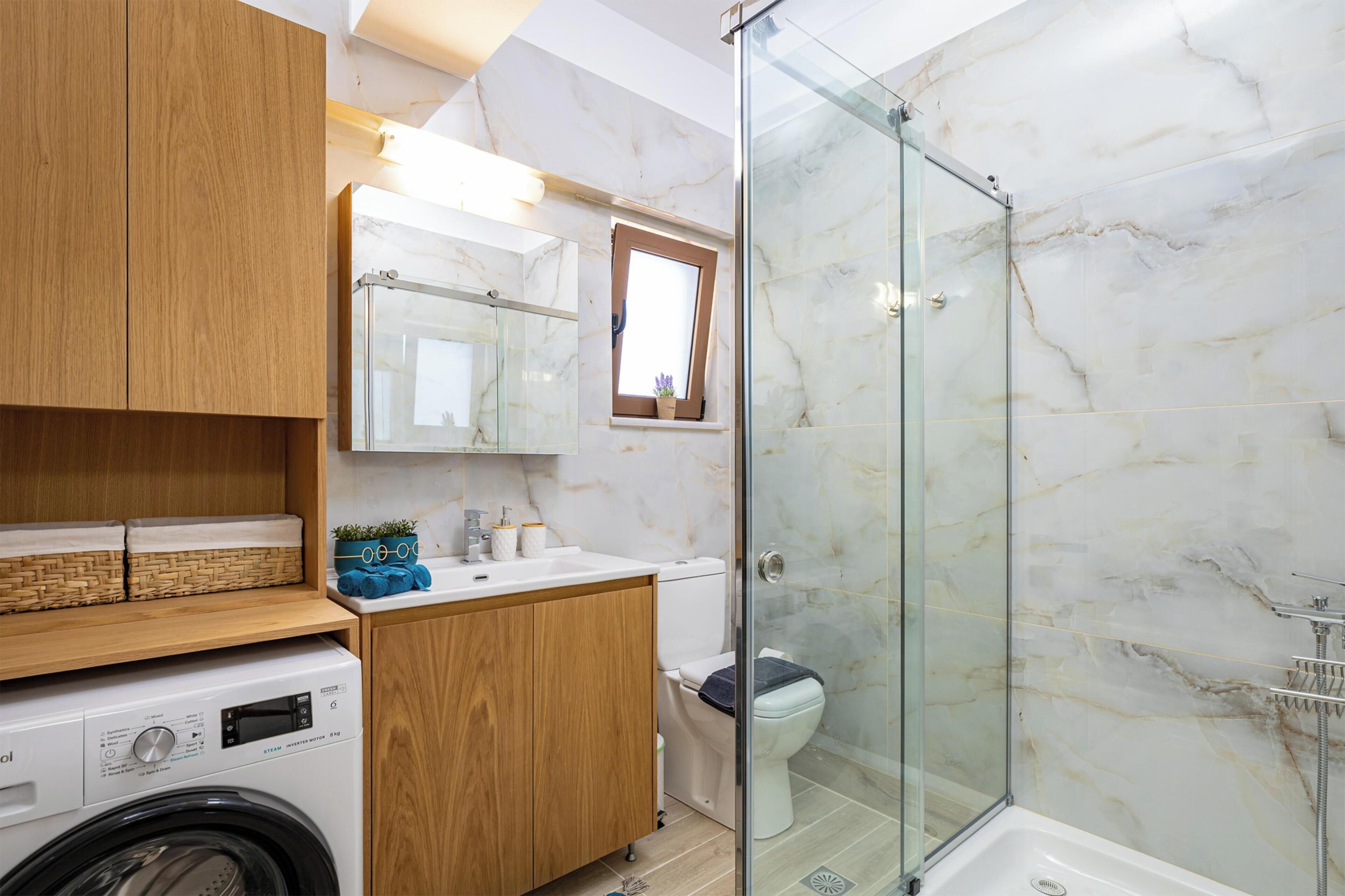 Een moderne badkamer met een douchecabine van glas, een ingebouwde wasmachine, houten kasten en een marmeren wand.