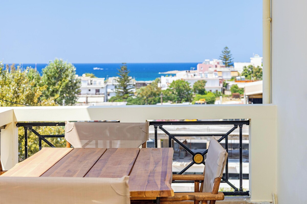 Ein Holztisch und Stühle auf einem Balkon mit Blick auf das Meer und Gebäude.