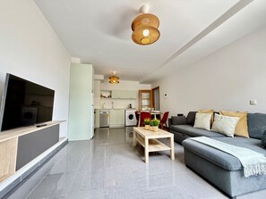 Ein modernes Wohnzimmer mit einem grauen Sofa, einem Holztisch und einem Fernseher auf einem Schrank. Im Hintergrund ist eine Küche mit weißen Schränken und einer Waschmaschine zu sehen.