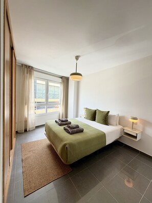 Ein modernes Hotelzimmer mit einem großen Bett, einem Nachttisch mit Lampe, Blick durch das Fenster und einem braunen Teppich.
