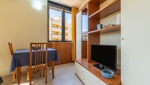 A room with a dining table, chairs, a TV on a wooden stand, and a window with curtains. - Accommodation with Private Parking - Cagliari (Cagliari)