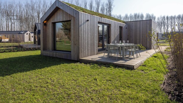 Una casa moderna de madera con un amplio ventanal, una terraza con mobiliario exterior y un jardín césped.