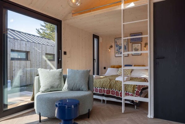 A modern bedroom with a sofa, a bed with patterned bedding, a blue stool, and a glass door leading outside.