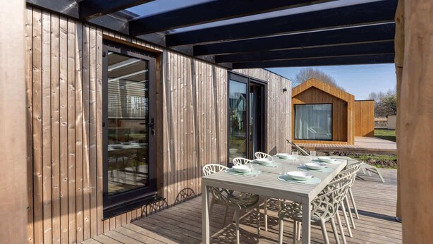 A wooden deck with a table set for dining, a glass door, and a view of a wooden structure in the background. - Tiny House with forest view in Oud Ade | Tuin van Keet - 27 (Oud Ade)