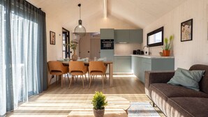A modern living room with a sofa, a dining table with chairs, a kitchen area, and a large window with curtains. - Tiny House with forest view in Oud Ade | Tuin van Keet - 27 (Oud Ade)