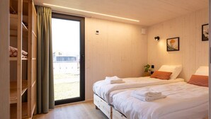 A bedroom with two beds, a wooden wardrobe, a sliding glass door, and a small plant on the wall. - Tiny House with forest view in Oud Ade | Tuin van Keet - 27 (Oud Ade)
