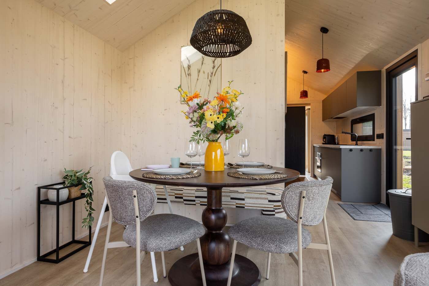A dining area with a round table, four chairs, a vase with flowers, and a kitchen in the background.