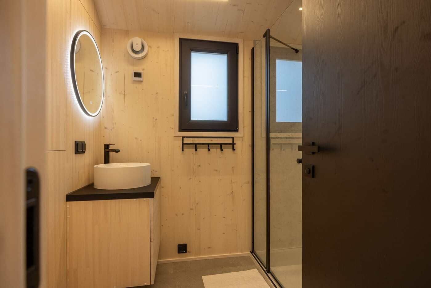 Un baño con un espejo redondo, un lavamanos, una ventana y una puerta de cristal para la ducha.