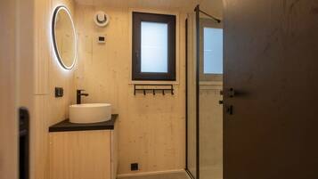Un baño con un espejo redondo, un lavamanos, una ventana y una puerta de cristal para la ducha.