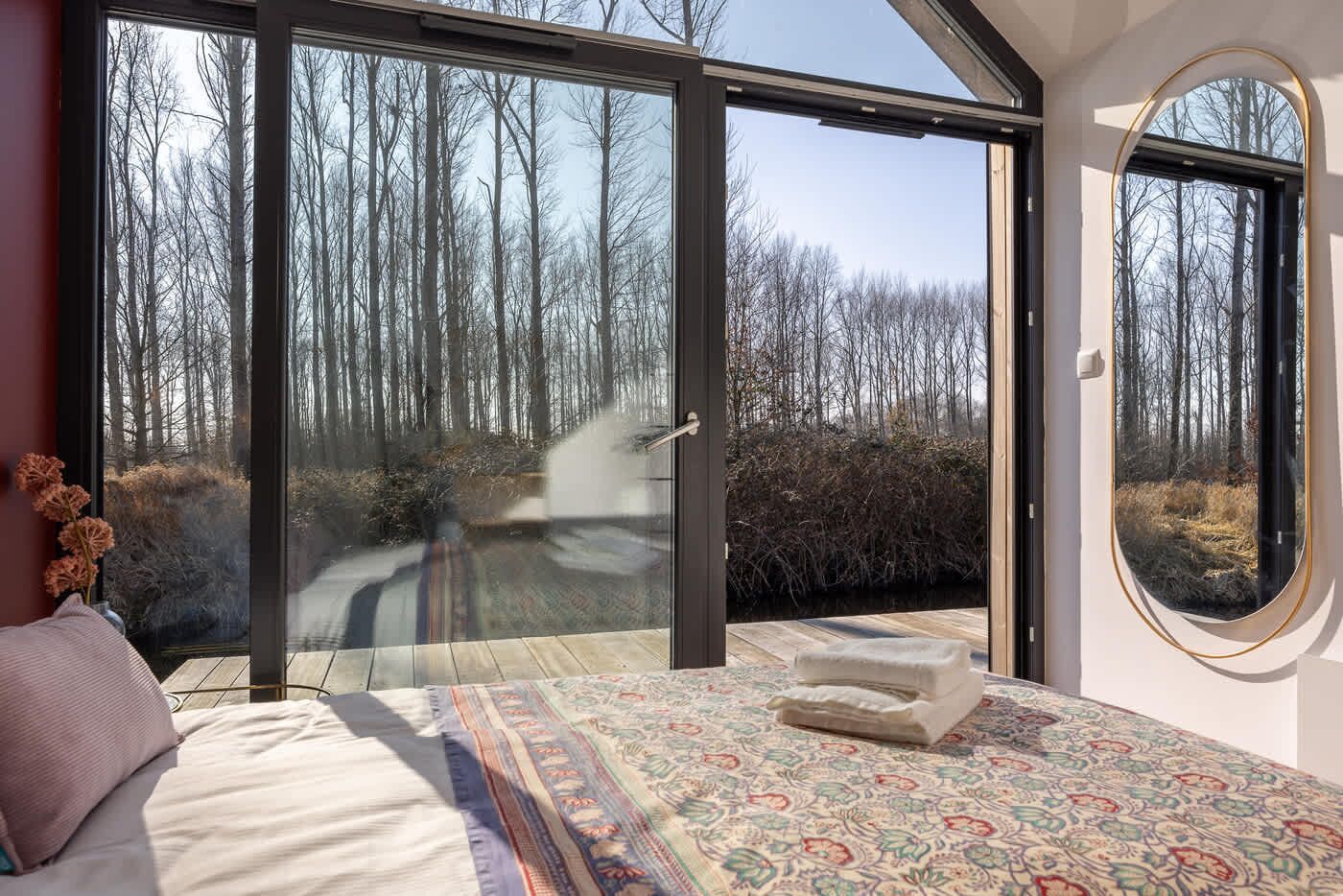 A bedroom with a large sliding glass door leading to a forest view, a bed with a patterned rug, and a small round window.