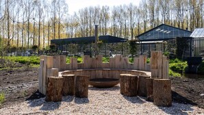 Un espace de détente circulaire en bois, doté d’un foyer, entouré d’un chemin de gravier, avec un bâtiment et des arbres en arrière-plan.