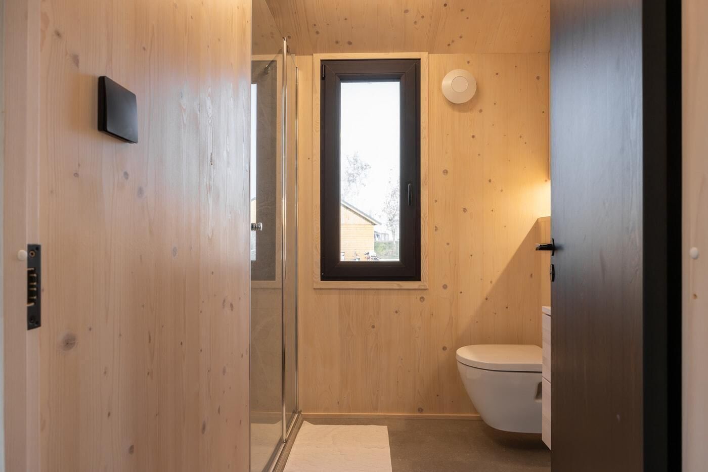 A wooden-paneled bathroom with a toilet, a window, and a shower.