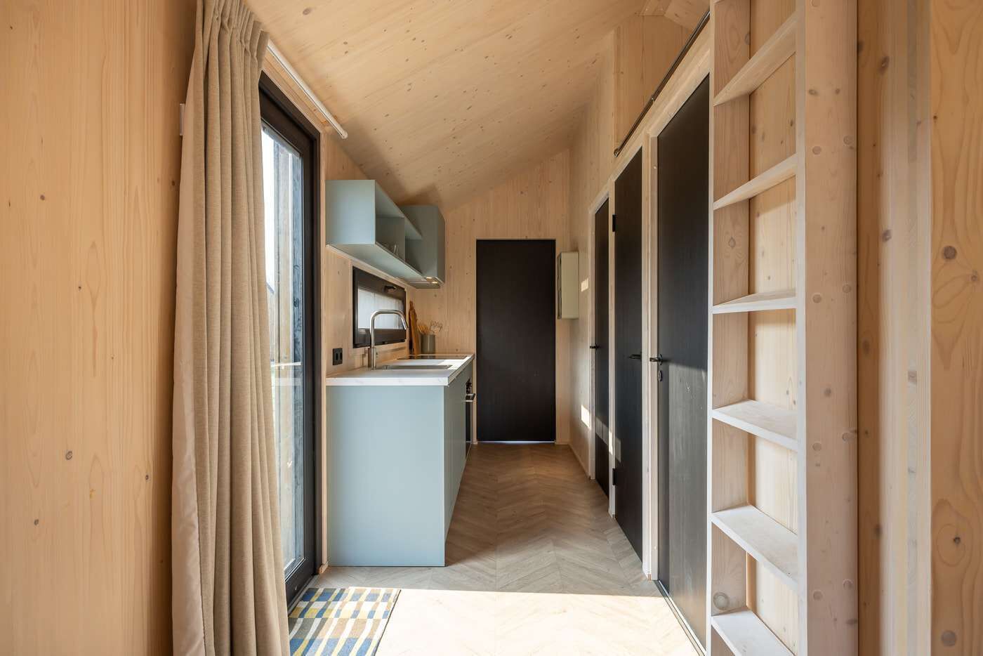 A narrow hallway with wooden walls, a kitchen area with a sink and cabinets, and a door leading to another room.