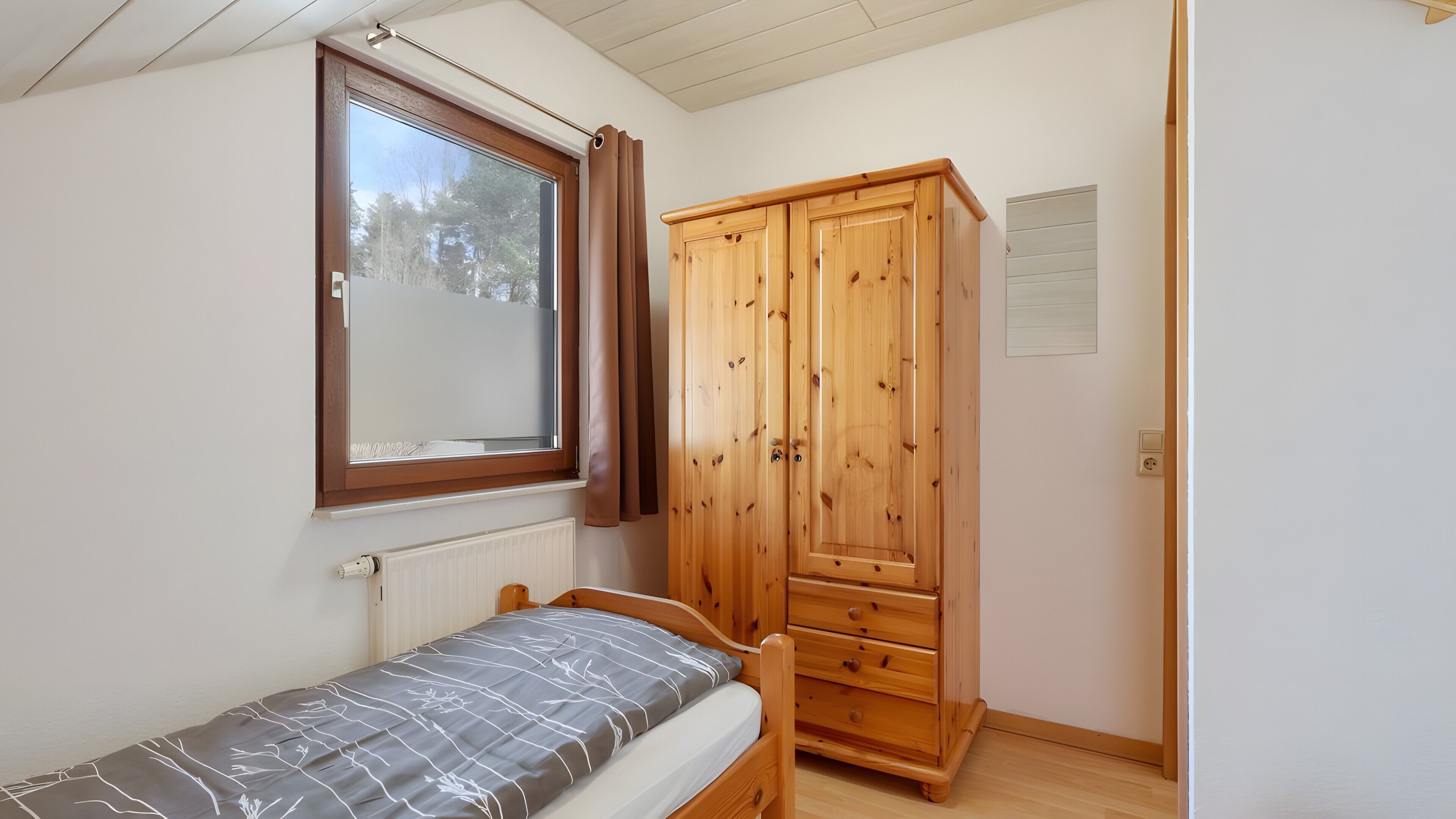 Ein Schlafzimmer mit einem Einzelbett, einem Holzschrank, einem Fenster mit Vorhängen und einer Heizung.