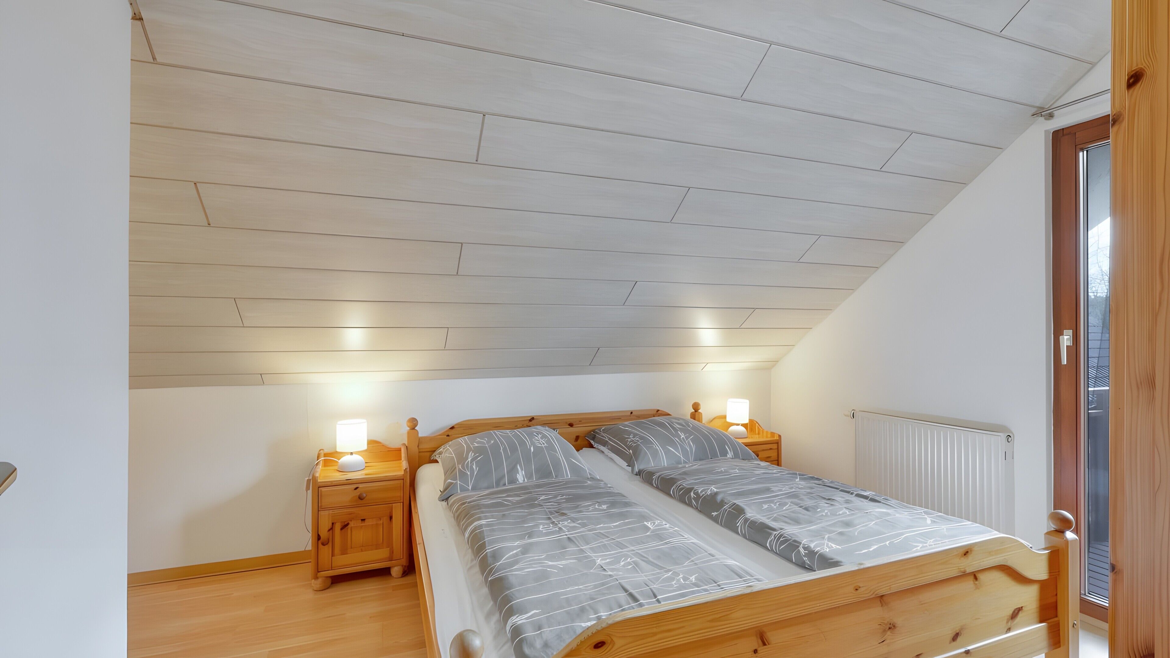 A bedroom with a sloped ceiling, wooden headboard, two bedside tables with lamps, a radiator, and a window.