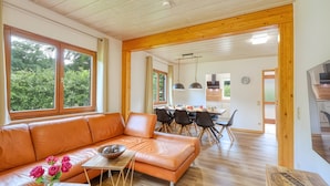 Un salon moderne avec un canapé en cuir, une table à manger et des chaises, des poutres en bois au plafond et de grandes fenêtres donnant sur un paysage verdoyant.