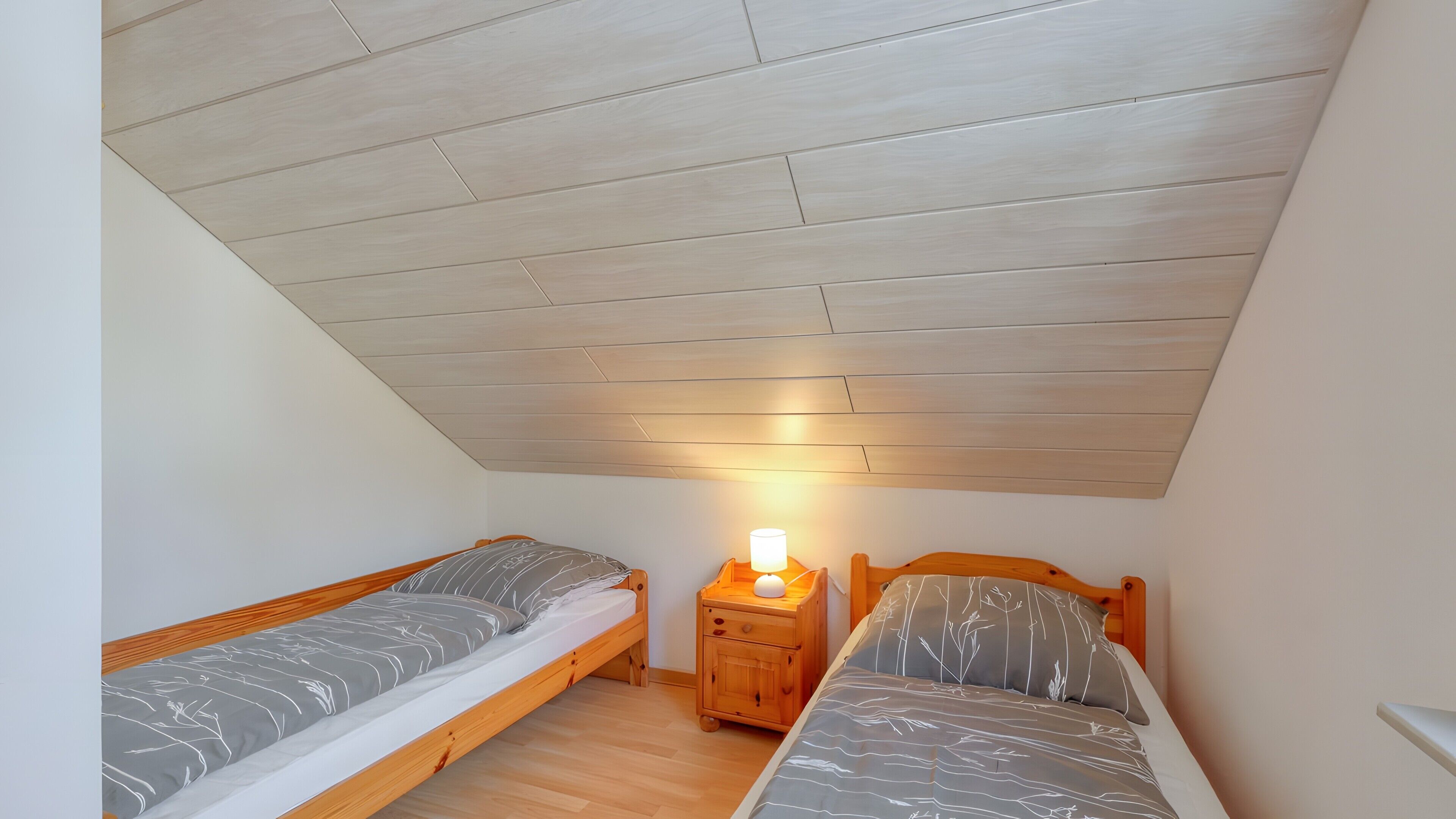 Een studentenkamer met twee houten stapelbedden, een nachtkastje met een lamp, en een schuine plafond.