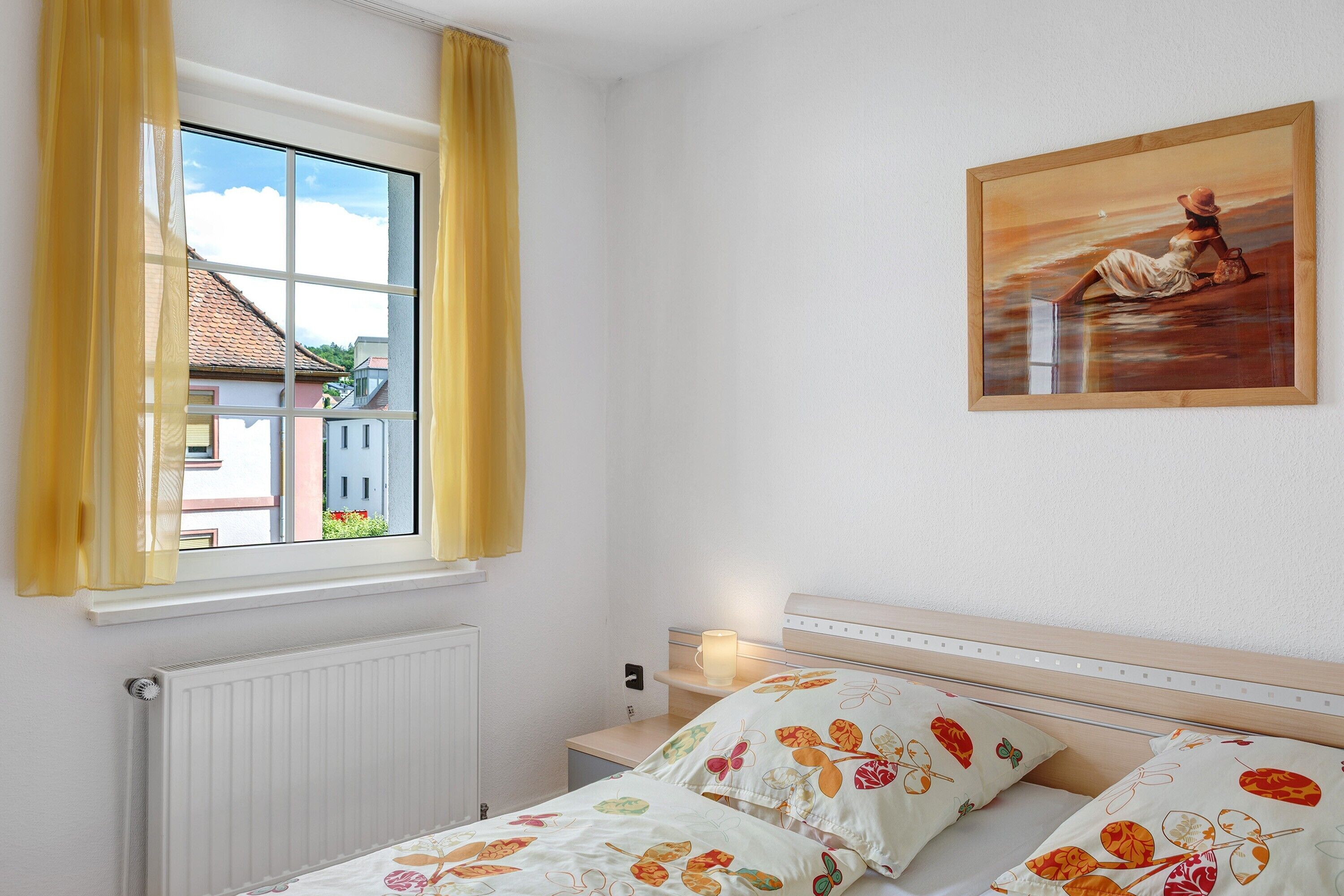 A bedroom with a large window, a bed with floral bedding, a nightstand with a lamp, and a painting on the wall.