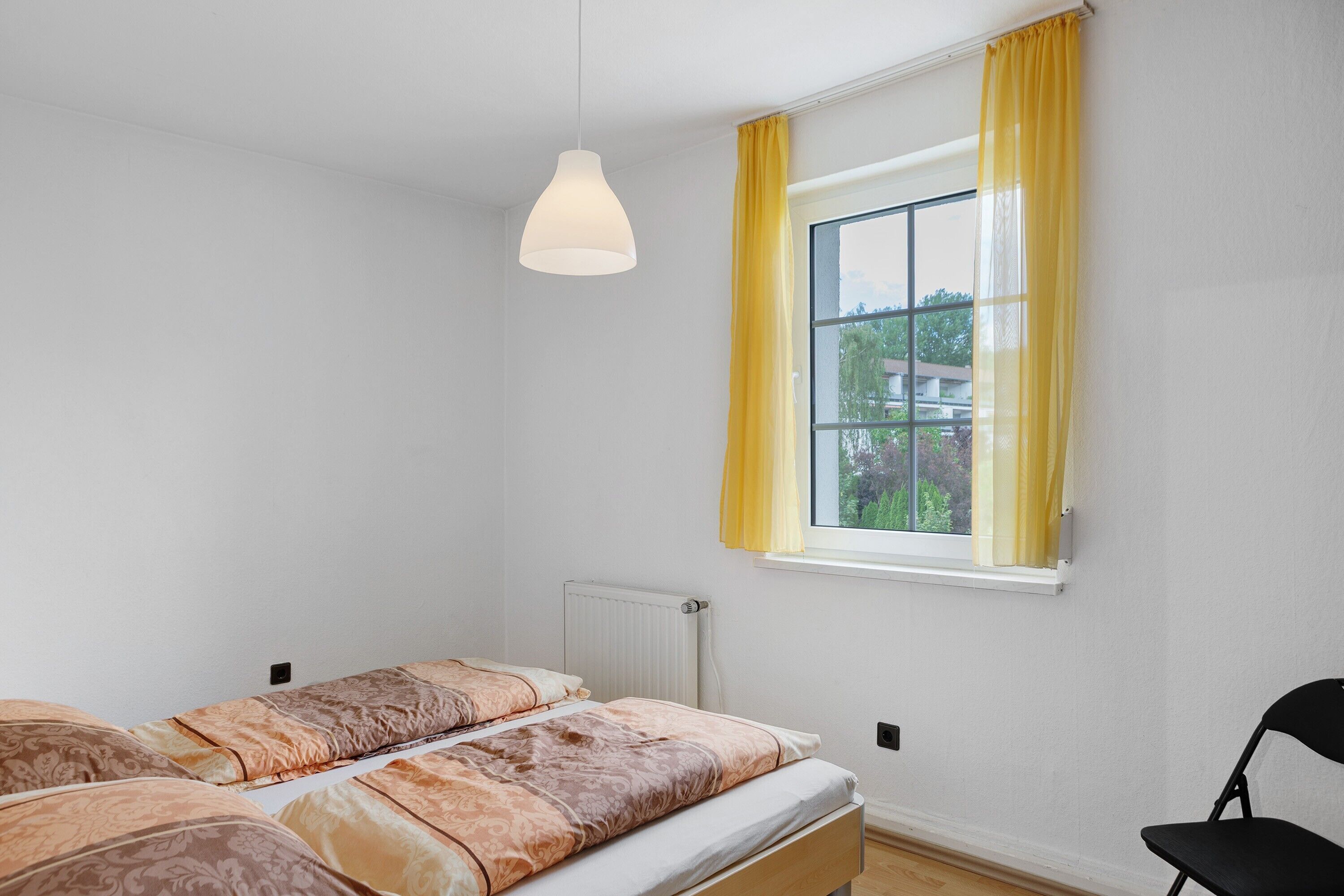Ein Schlafzimmer mit einem Bett, einem Sessel, einem Fenster mit Vorhängen und einer Pendelleuchte.