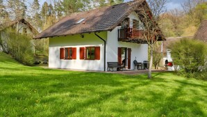 Une maison à deux étages avec un toit en pente, une terrasse avec une table et des chaises, et une pelouse verdoyante.