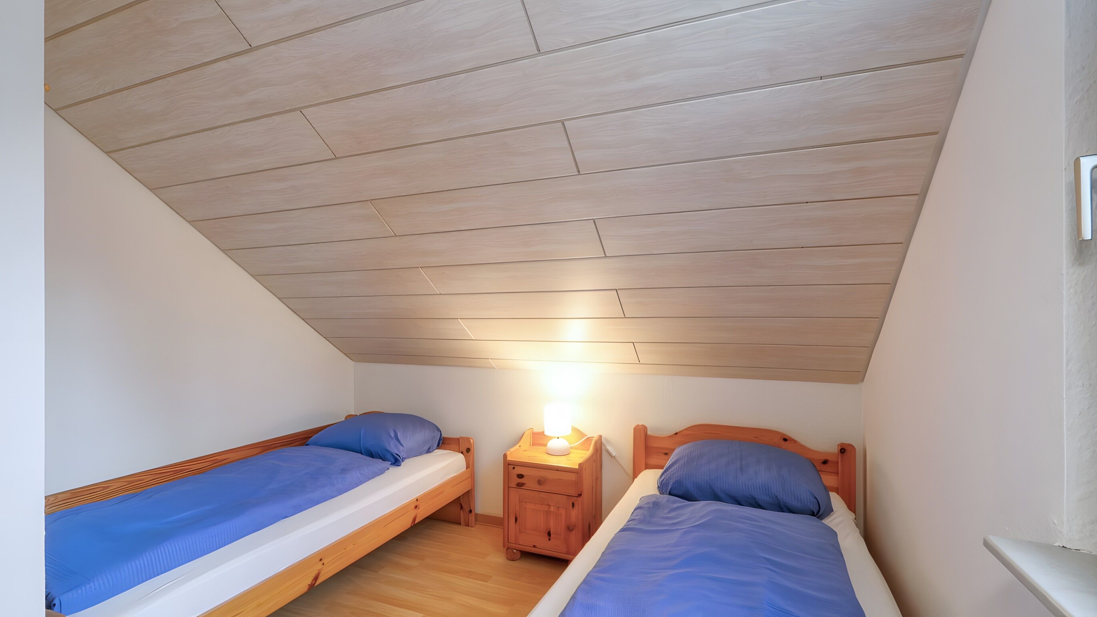 A small, single-room accommodation with two wooden bunk beds, a nightstand with a lamp, and a sloped ceiling.