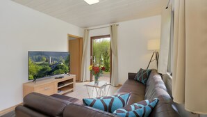 Un salon avec un téléviseur, un canapé et une vue sur la verdure, grâce à une porte ouverte.