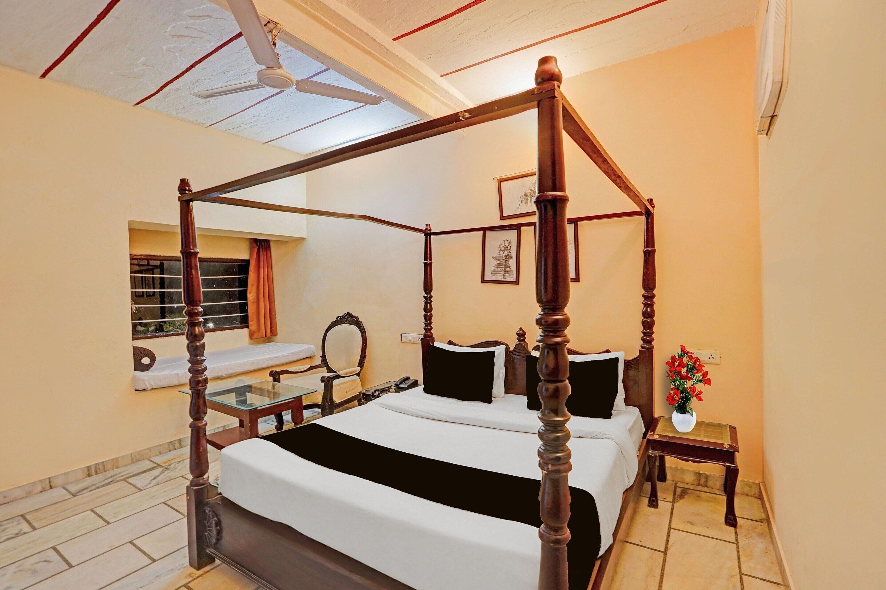 A four-poster bed with a white bedspread, a small bedside table with a vase of flowers, and a window with curtains.
