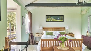 Une chambre à coucher avec un lit, des tables de chevet, une commode, un banc en bois, une petite table avec un vase et une fenêtre avec des rideaux.