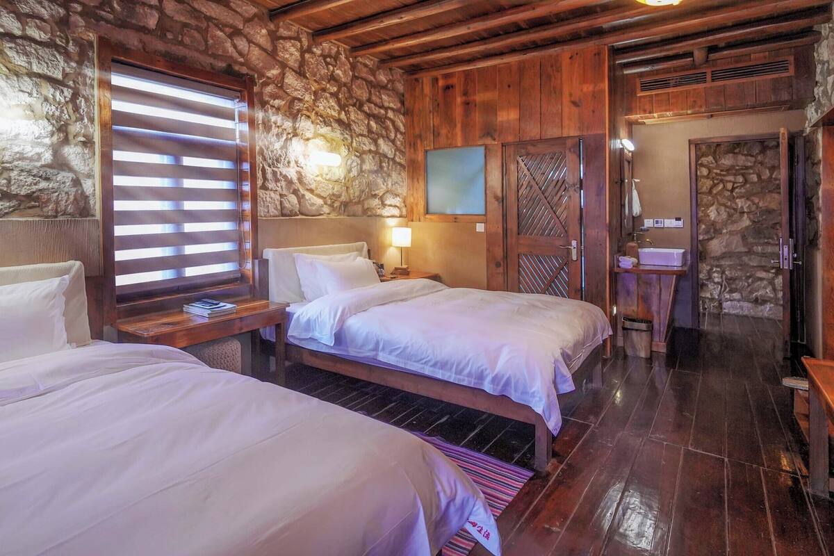 Une chambre avec deux lits, des murs en pierre, des poutres en bois, une cheminée en pierre et une porte en bois.