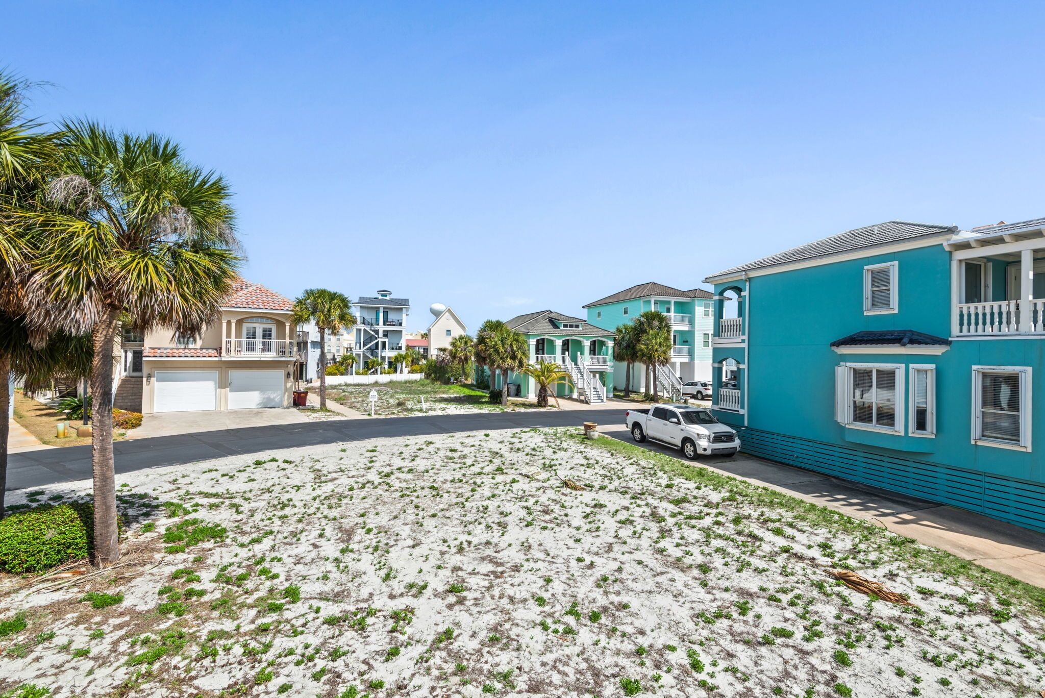 A residential street with houses, a white car parked on the street, and a sandy area with sparse vegetation.