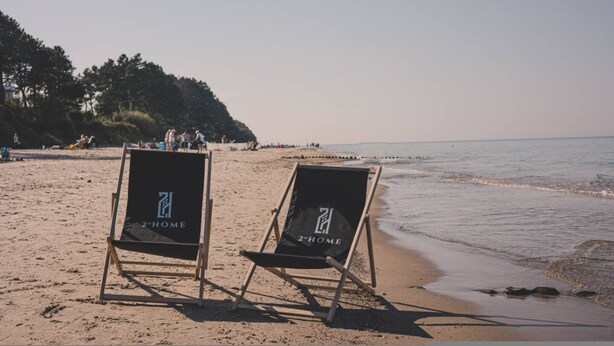 Två strandstolar med logotypen ”2-HOME” placerade på en sandstrand, med ett lugnt hav och en skogbeklädd kulle i bakgrunden.