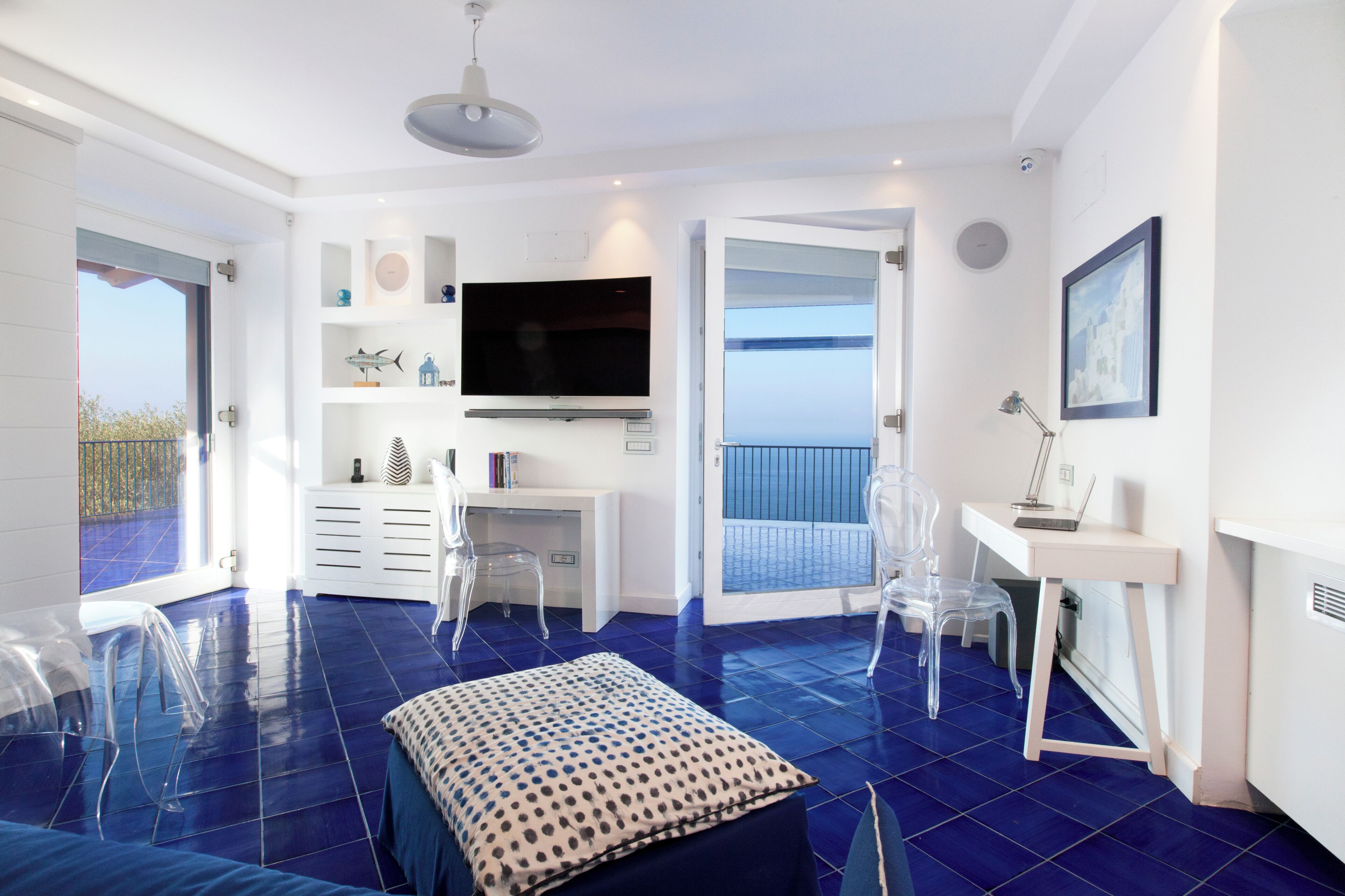Quarto moderno com uma televisão grande, cadeiras de plástico transparente, uma escrivaninha e vista para o mar.