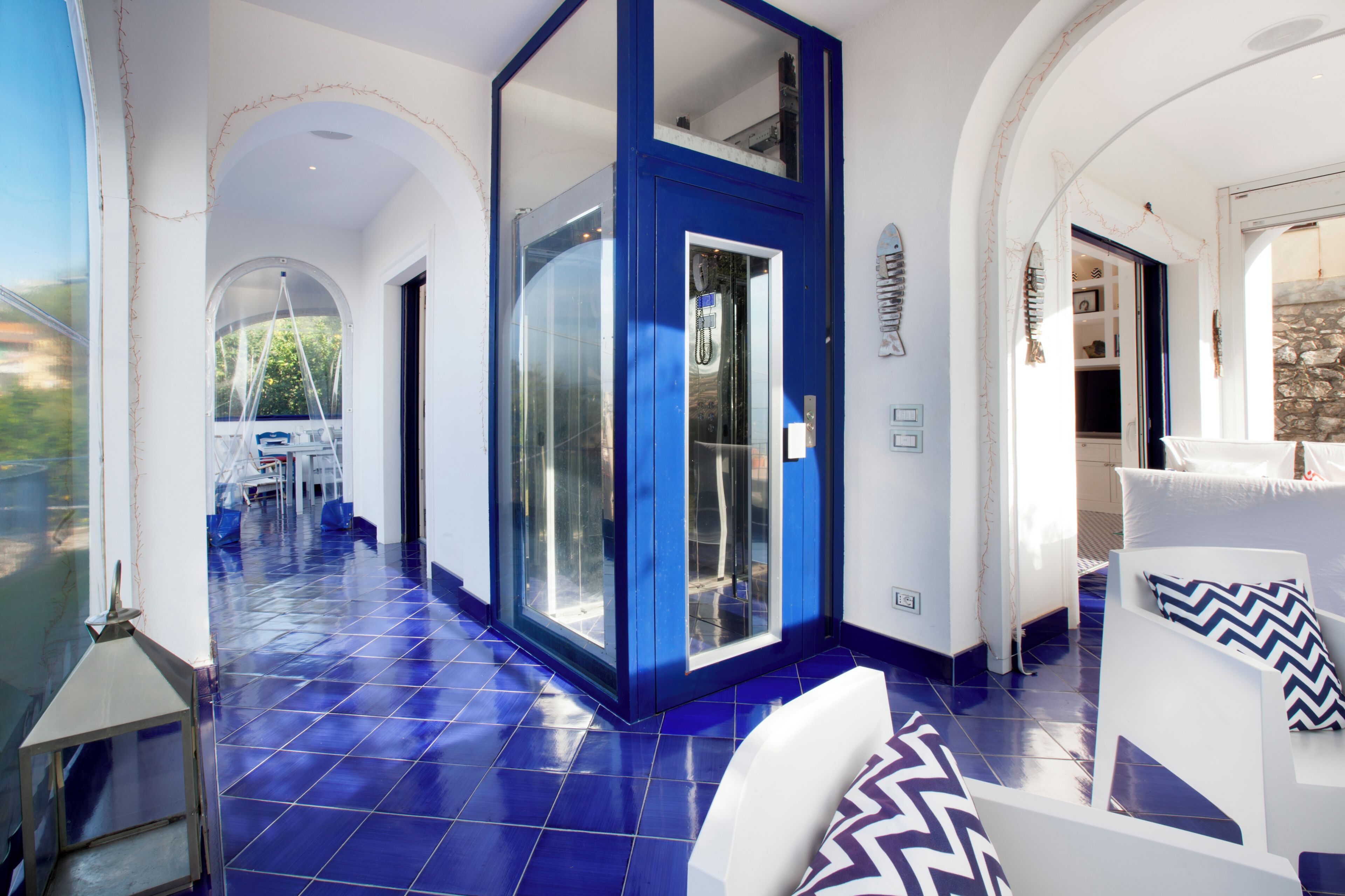 Um corredor espaçoso com parede azul decorativa, porta de vidro e sofá branco com almofadas estampadas.