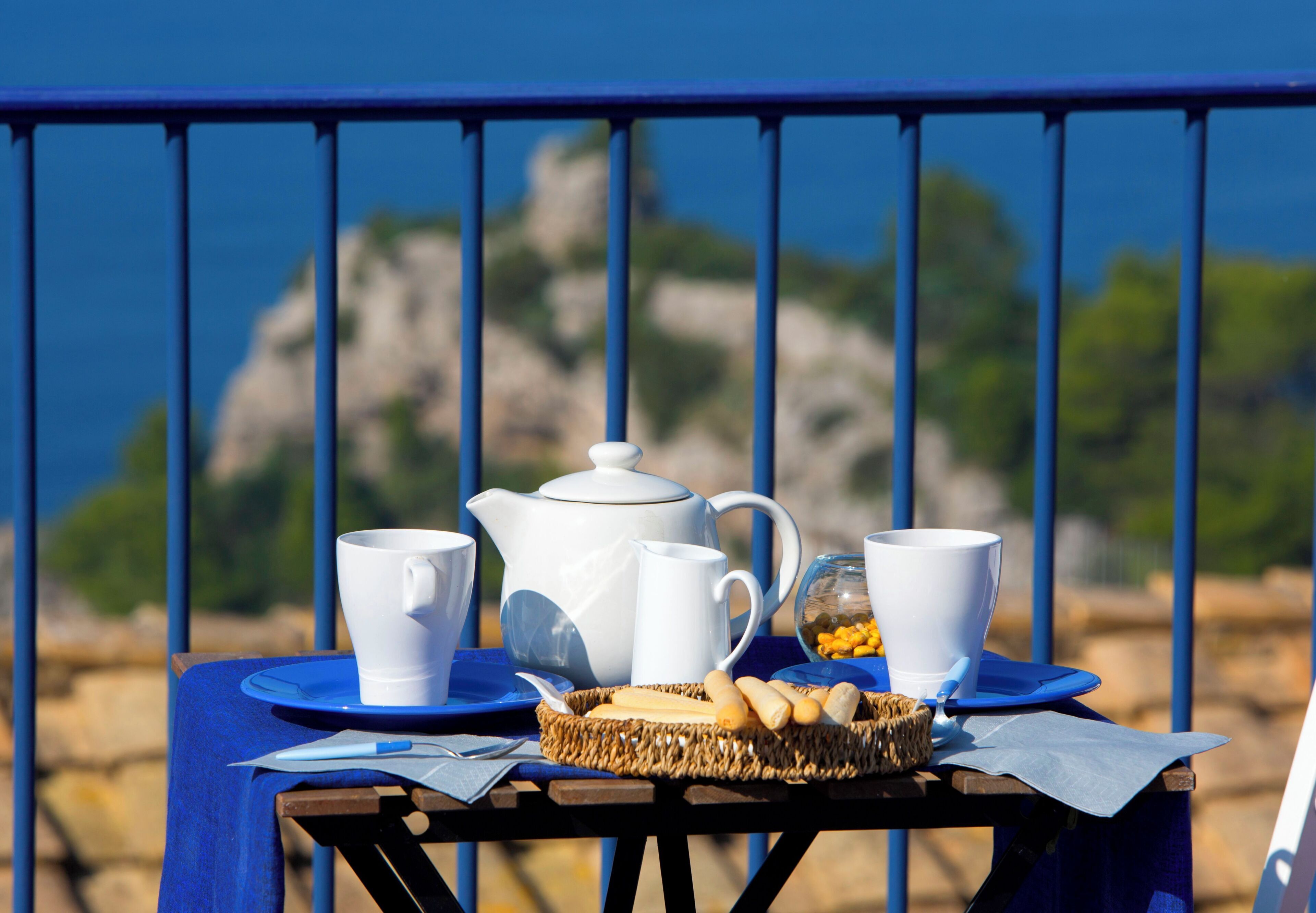 Mesa com uma chaleira branca, duas chávenas brancas, um prato azul e uma cesta de pão, apoiada sobre uma mesa de madeira com toalha de mesa azul, com uma grade azul e uma paisagem rochosa ao fundo.