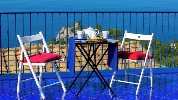 A balcony with two white chairs, a table with a blue tablecloth, and a view of the sea. - De Vivo Realty - Villa Blue Skyline (Marciano)