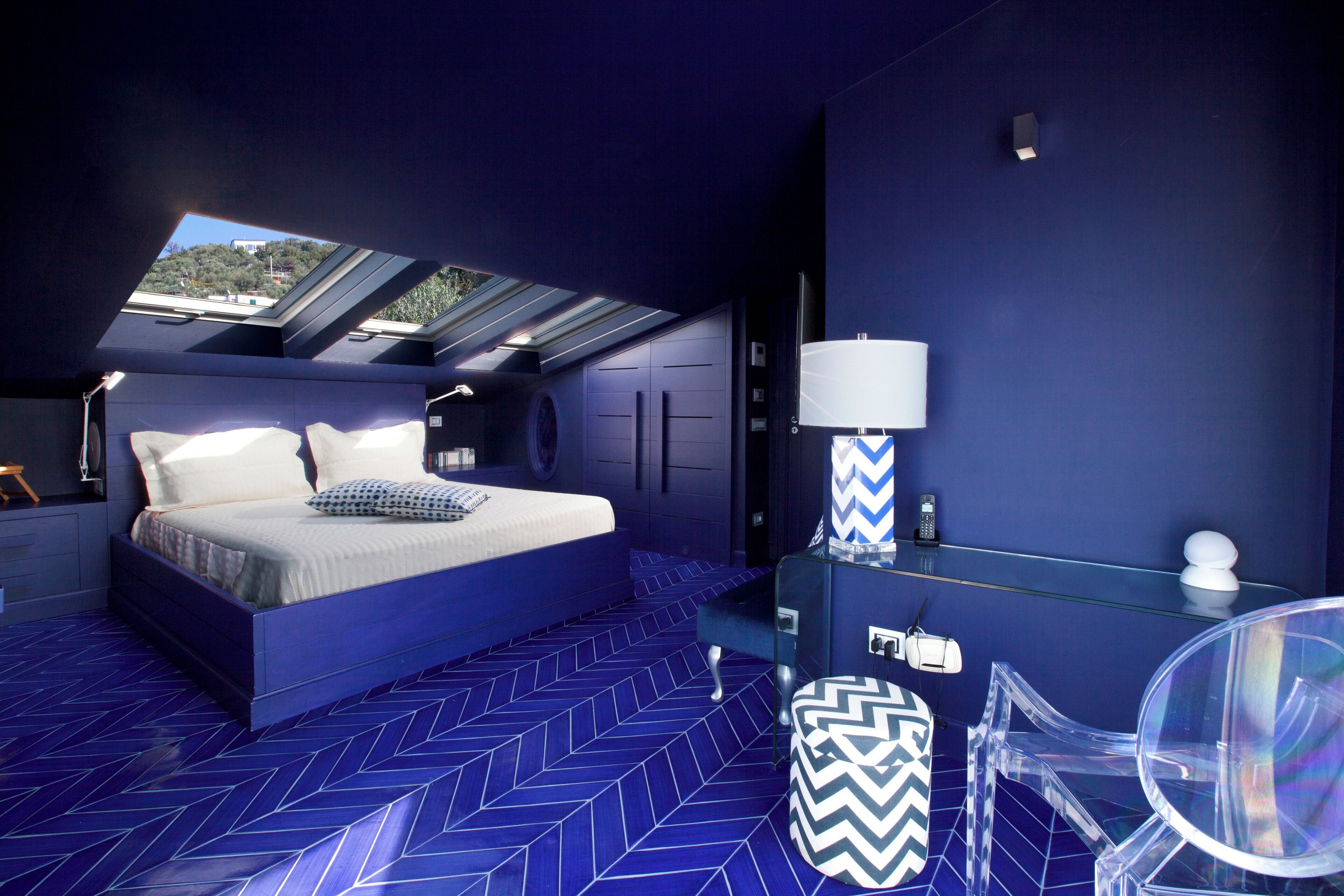 A bedroom with a skylight, a bed with pillows, a bedside table with a lamp, and a clear glass chair.