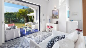 A modern hotel room with a large sliding glass door leading to a garden, a bed with patterned bedding, a desk with a laptop, and a small seating area. - De Vivo Realty - Villa Blue Skyline (Marciano)