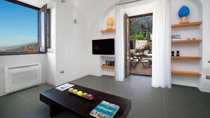 Ein modernes Wohnzimmer mit einem Flachbildfernseher, einem Couchtisch mit einem Buch und farbigen Gegenständen, einem Kamin und Blick durch ein offenes Fenster.