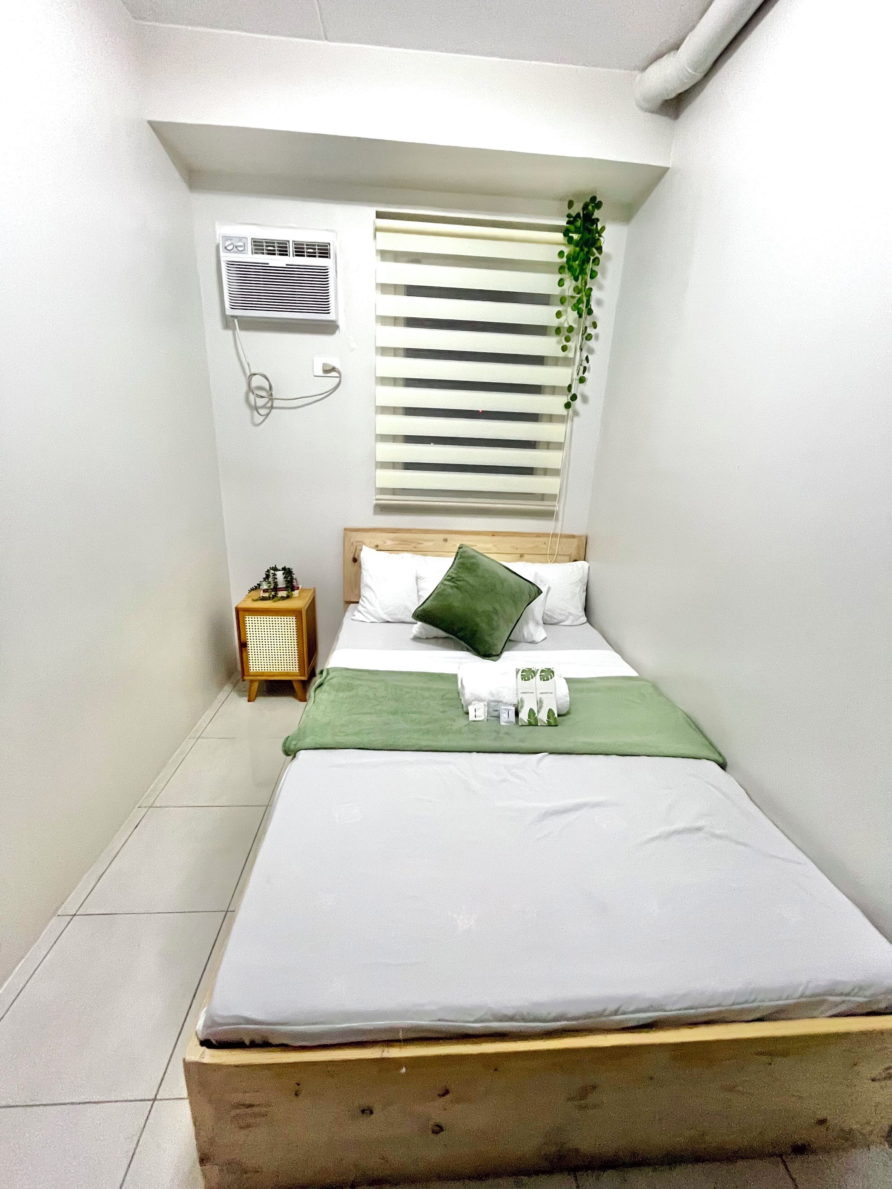 Una habitación pequeña y limpia con una cama, una mesita de noche, aire acondicionado instalado en la pared y una ventana con persianas.