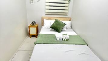 Una habitación pequeña y limpia con una cama, una mesita de noche, aire acondicionado instalado en la pared y una ventana con persianas.