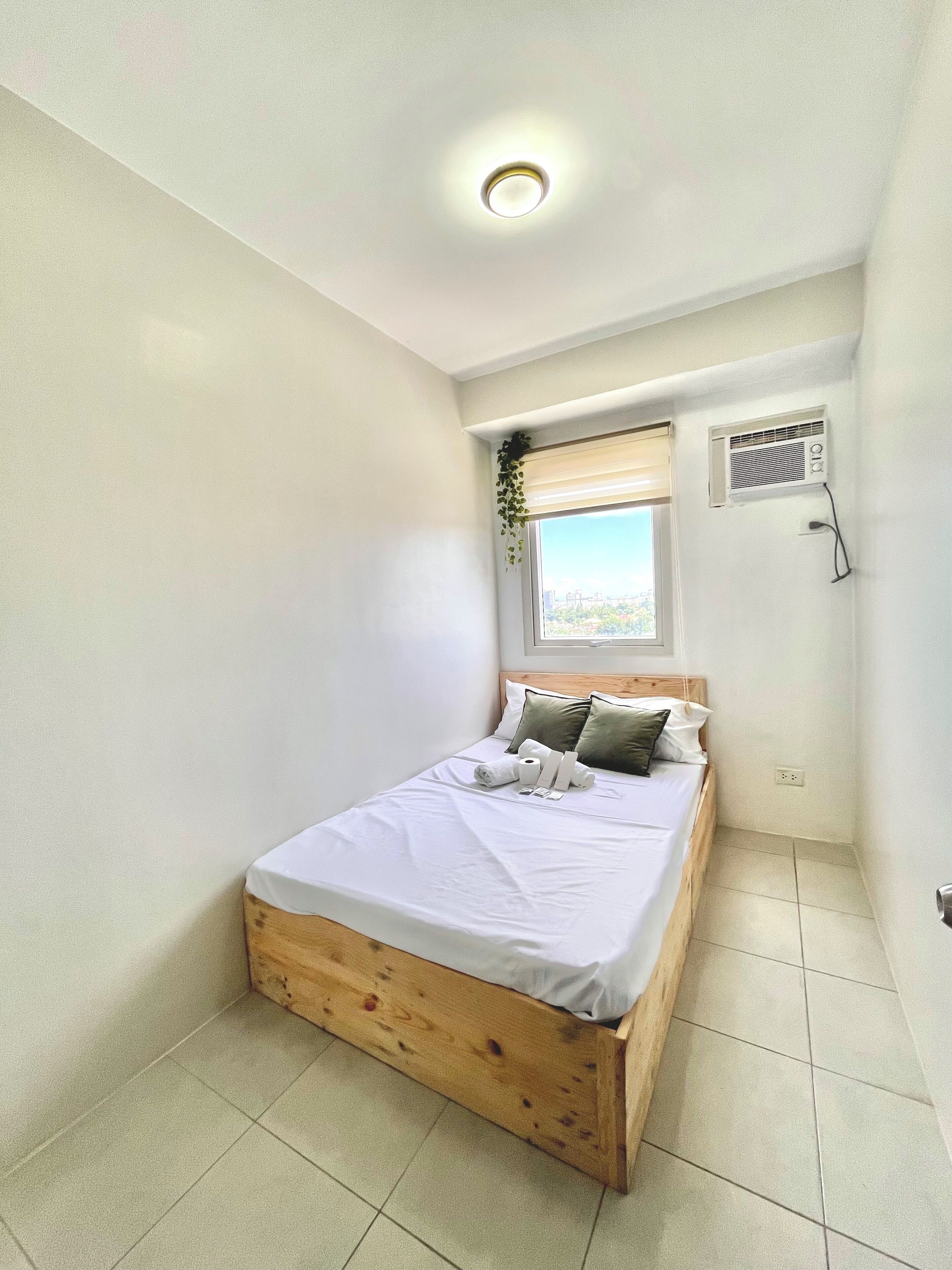 A small, neatly arranged bedroom with a single bed, a window with blinds, an air conditioning unit, and a ceiling light.