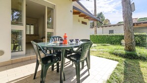 An outdoor patio with a table and chairs, a bottle of orange juice, and a view of a garden and neighboring buildings. - Charming Semi-detached Villa in Lignano (Lignano Sabbiadoro)