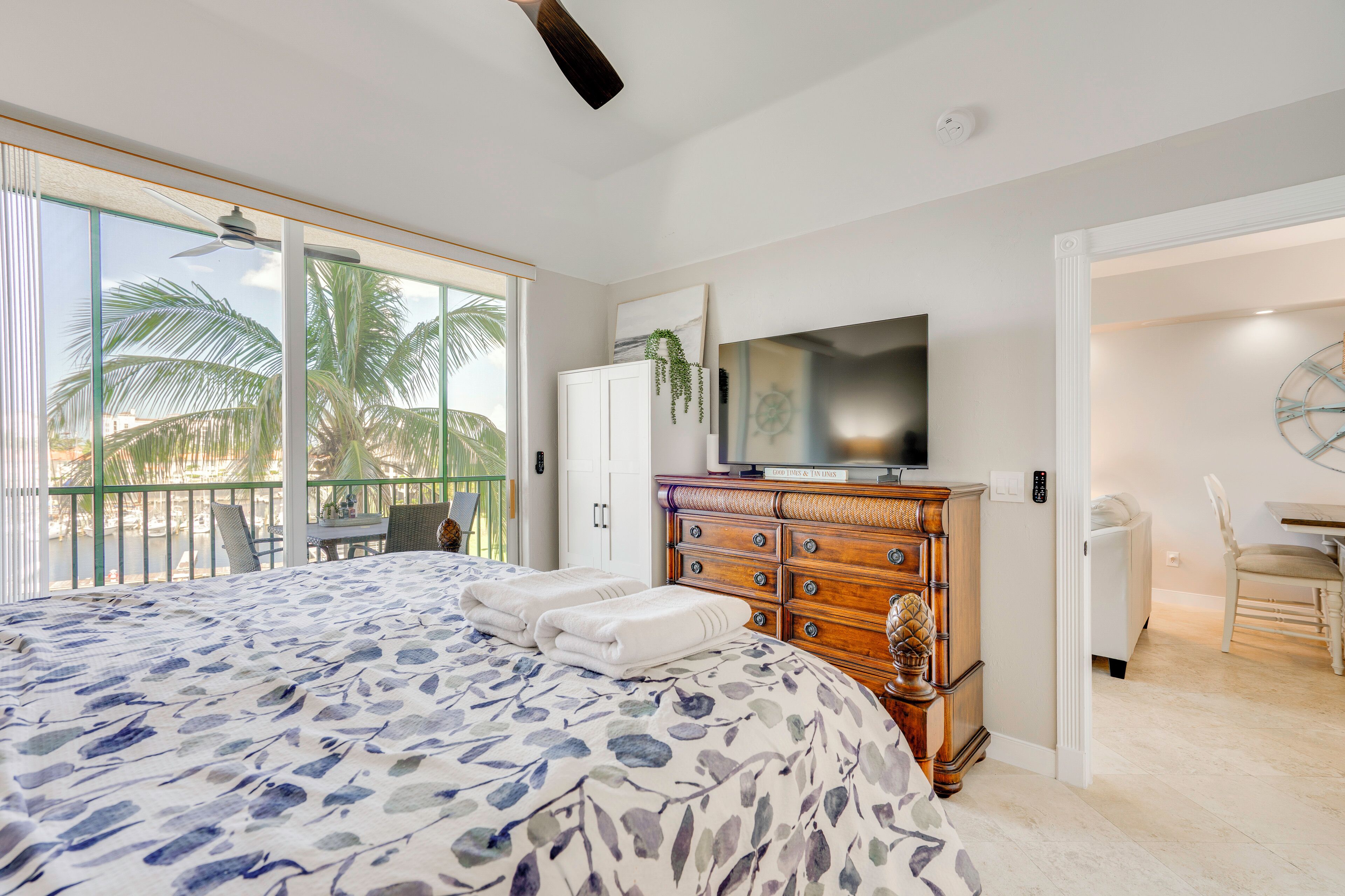 Un dormitorio con cama, cómoda, televisor y balcón con palmeras.