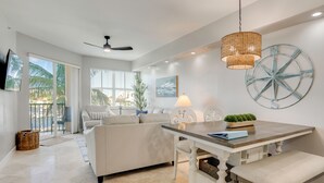 Uma sala de estar espaçosa com um sofá branco, uma mesa de jantar com um banco, um ventilador de teto e uma grande janela com vista para palmeiras.