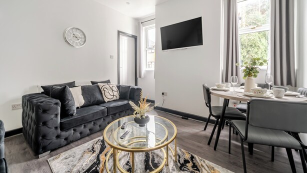 Een moderne woonkamer met een zwarte bank, een glazen salontafel, een eettafel met stoelen, een flatscreen televisie en een raam met gordijnen.