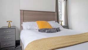 Een netjes opgemaakt bed met hoofdbord, een nachtkastje met een lamp, en een raam met gordijnen.