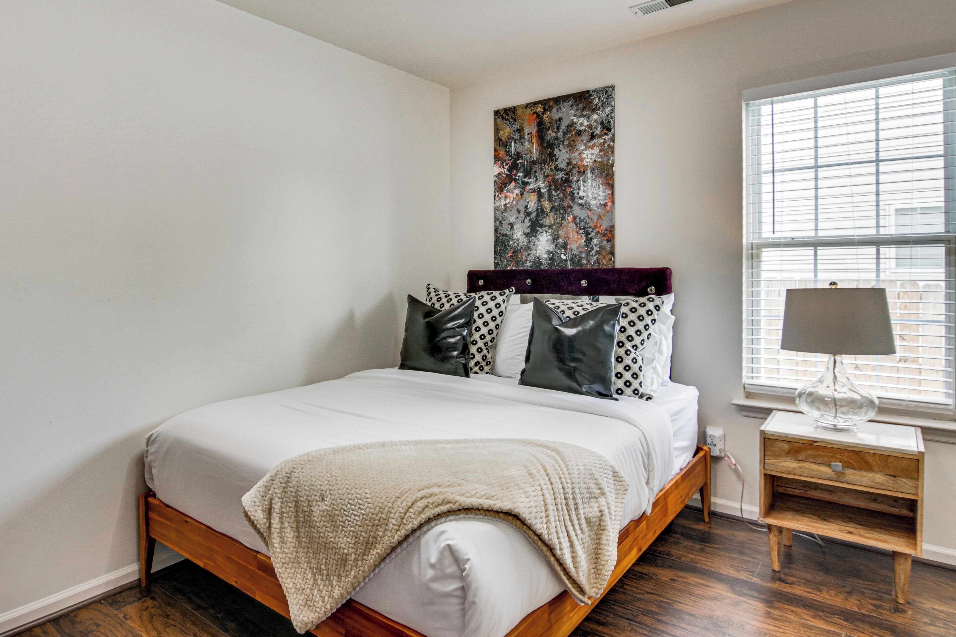 A bedroom with a bed, a nightstand, a lamp, a window with blinds, and a wall art piece.
