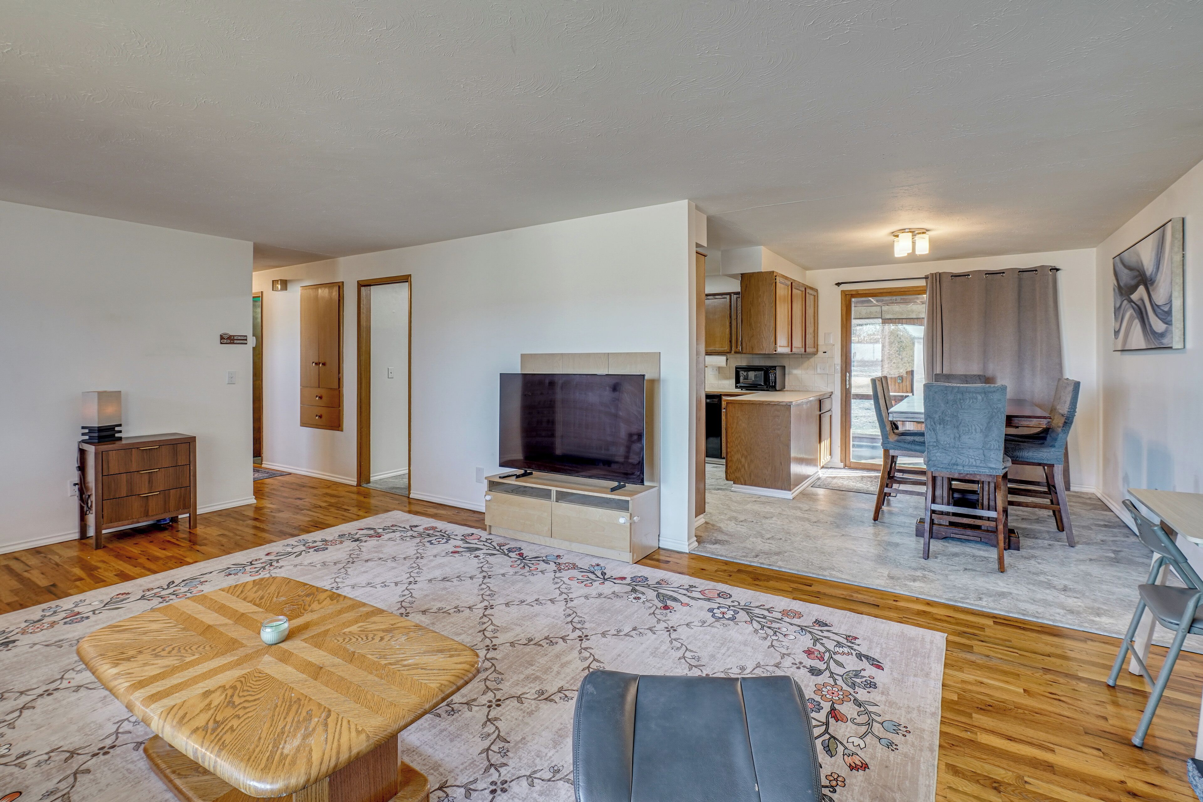 A spacious living area with a flat-screen TV, wooden furniture, a dining table with chairs, and a kitchen in the background.
