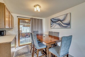 Un coin repas avec une table en bois, des chaises à l’assise rembourrée de couleur bleue, un tableau accroché au mur, et un espace cuisine doté de meubles en bois.