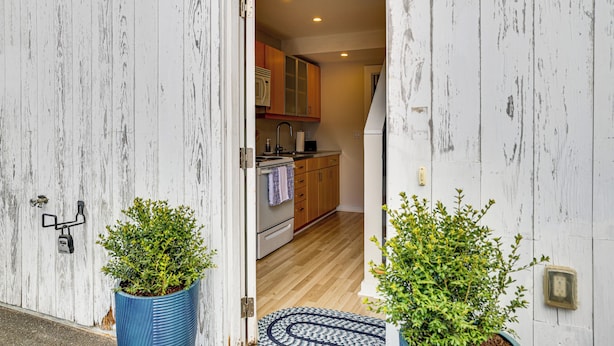 Une porte donne sur une cuisine équipée de meubles en bois, d’une cuisinière blanche et d’un tapis bleu sur le sol.